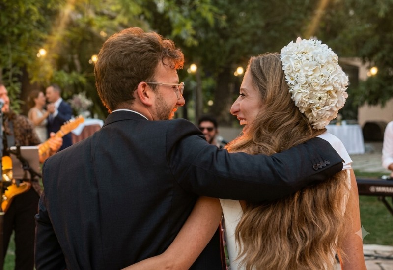 Texto Alternativo (Alt Text): Novios de espaldas, abrazados y sonrientes, mirando un concierto de música en directo durante su boda al aire libre en un jardín al atardecer, con la banda El Bueno, El Feo y El Malo tocando de fondo.