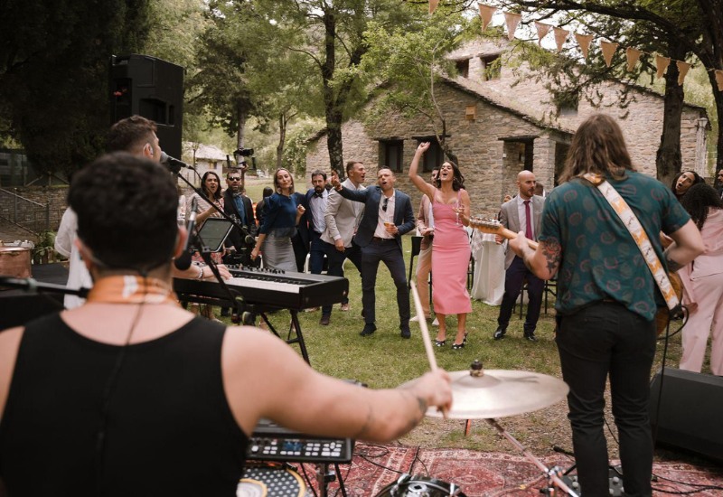 El grupo de versiones El Bueno, El Feo y El Malo actuando en directo sobre un escenario exterior durante un festival de verano, con el público bailando y luces de escenario profesionales bajo un cielo azul claro